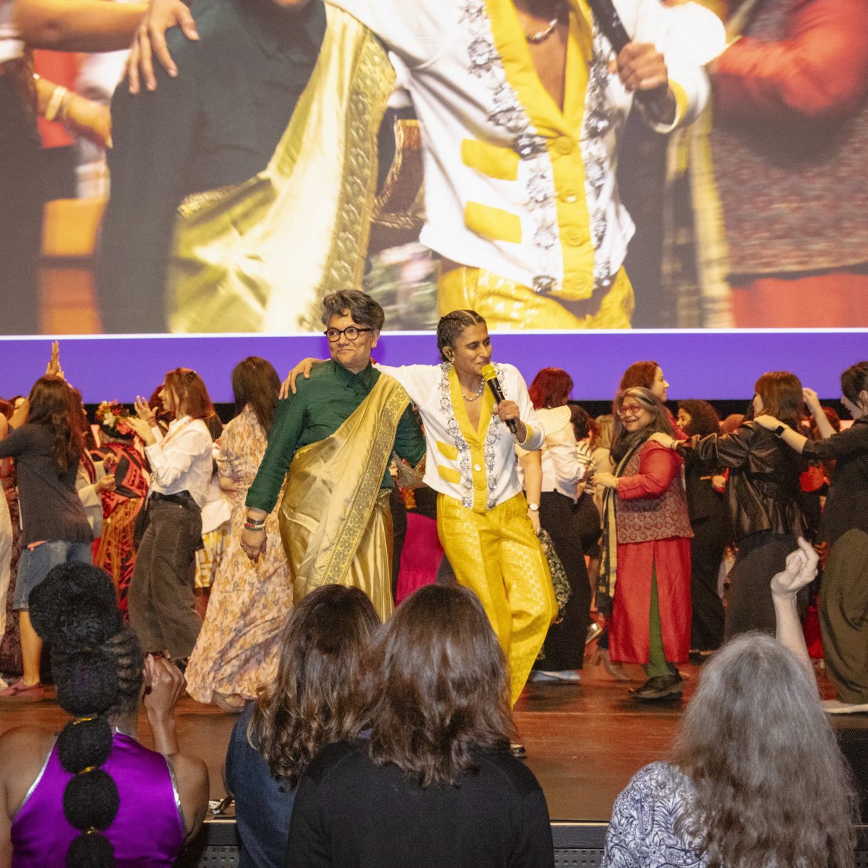 Women dancing on a stage, with other women watching as spectators in the foreground