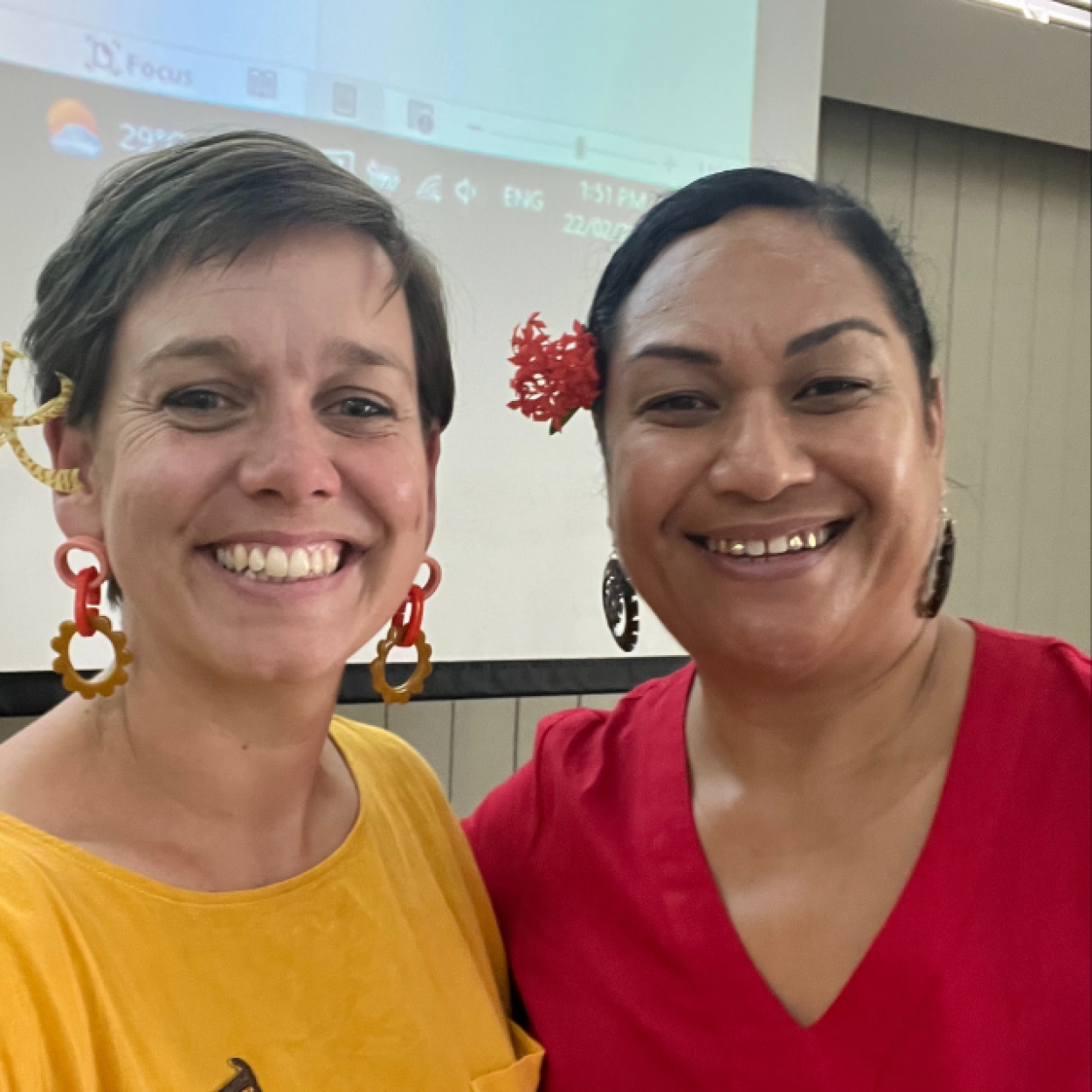 Two people smiling together. One has short hair and a yellow shirt, the other person has a red shirt and red flower behind their ear