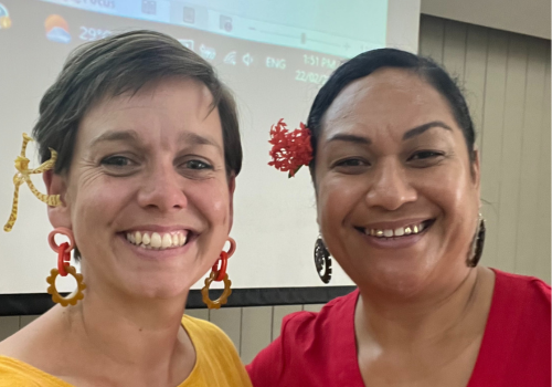 Two people smiling together. One has short hair and a yellow shirt, the other person has a red shirt and red flower behind their ear
