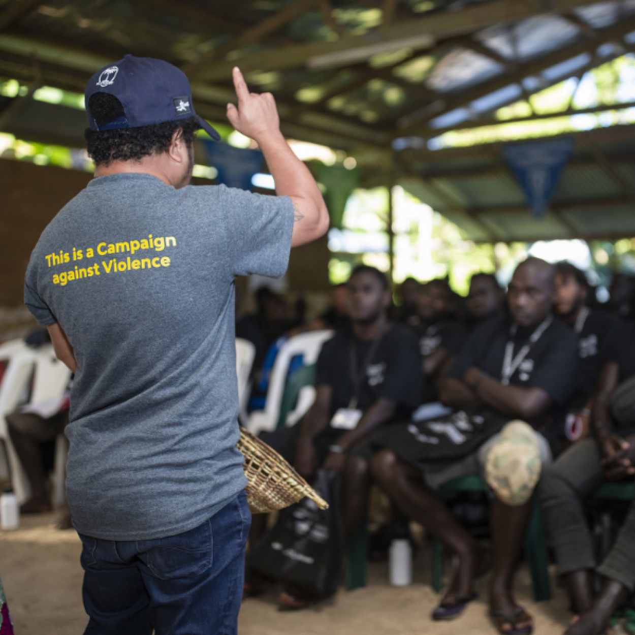 A man stands in front of an audience of men with his back to the camera. "This is a campaign against violence is written on the back of his shirt"