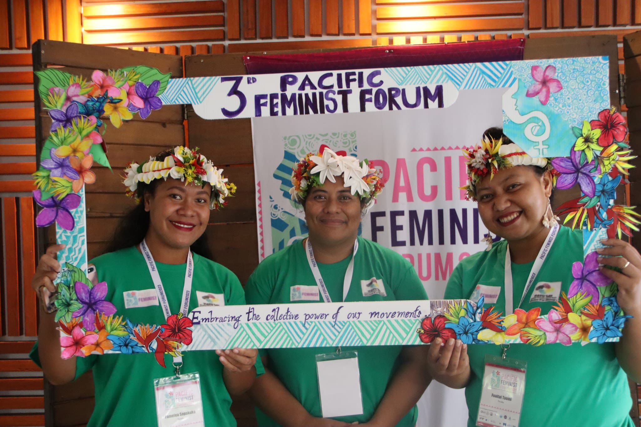Three women are smiling at the camera as they pose with a selfie frame around which the words '3rd Pacific Feminist Forum: Embracing the collective power of movements' are written. The frame is decorated with hand-drawn flowers from the Pacific against a backdrop of water.
