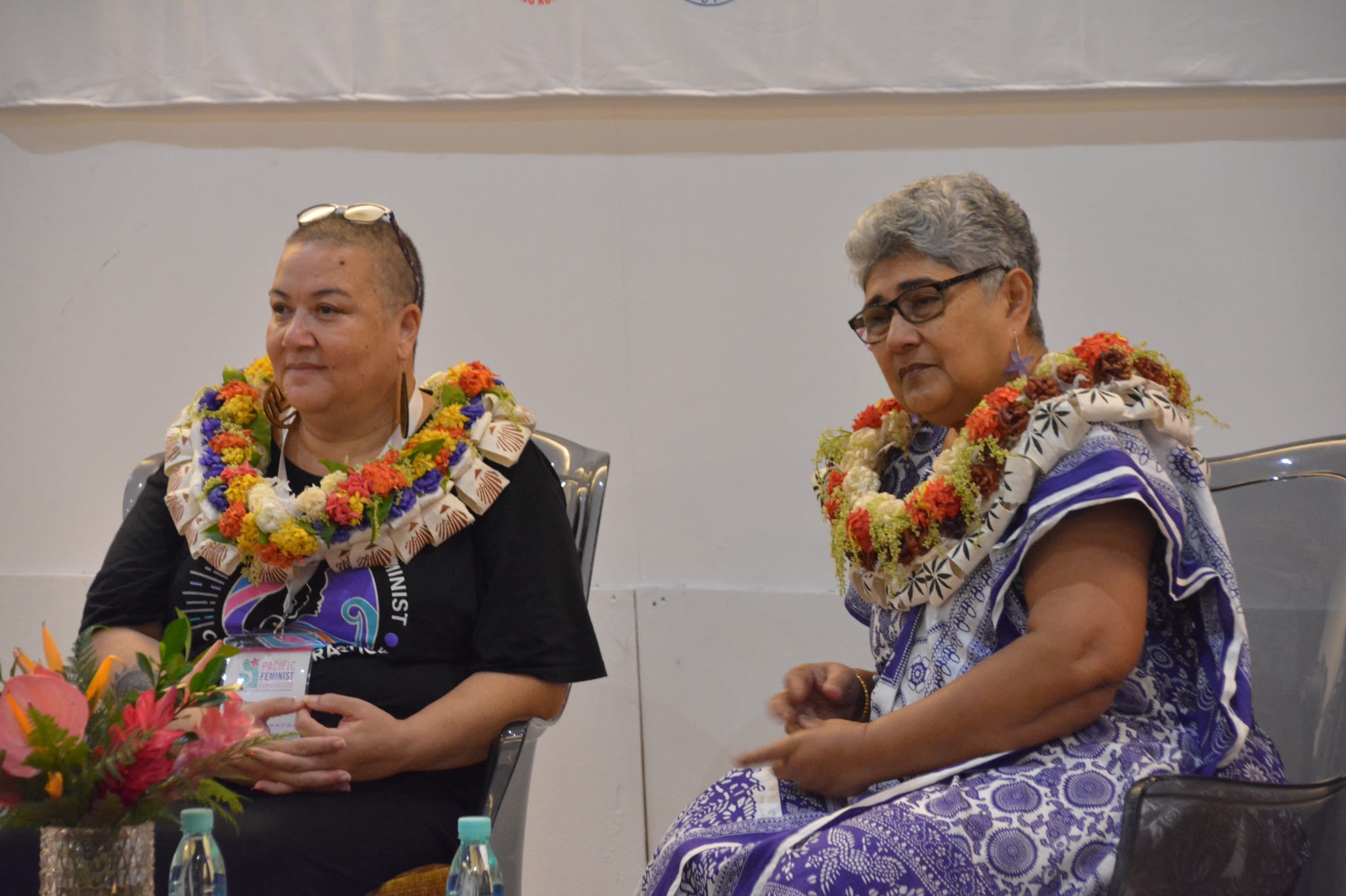 Noelene Nabulivou and Shamima Ali are sitting together on stage as they look towards the crowd. Both are wearing salusalus around their necks.