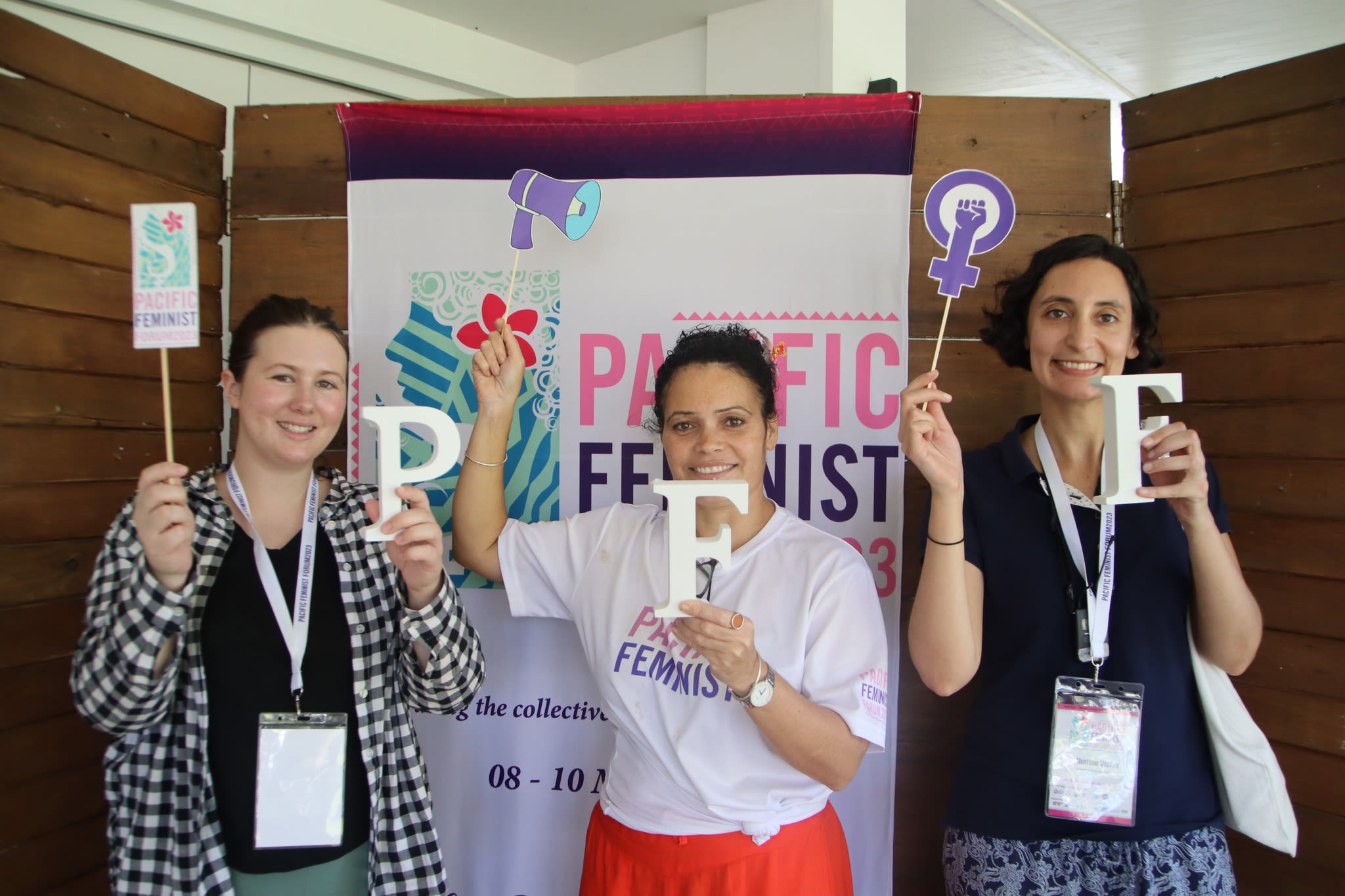 Three women are posing together in front of a banner that reads 'Pacific Feminist Forum 2023'. They are holding up letters that together spell out 'PFF'.