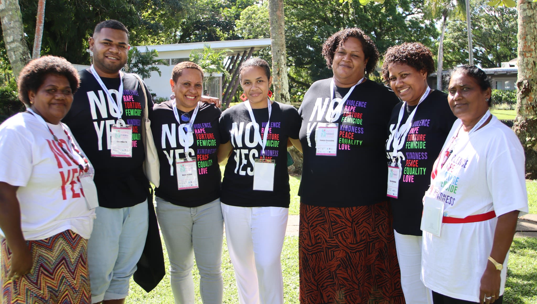 A group of men and women are posing together for the camera. It is daytime and they are standing in an outdoor area. They are wearing t-shirts that read 'No violence, rape, torture, femicide. Yes kindness, peace, equality, love.' 